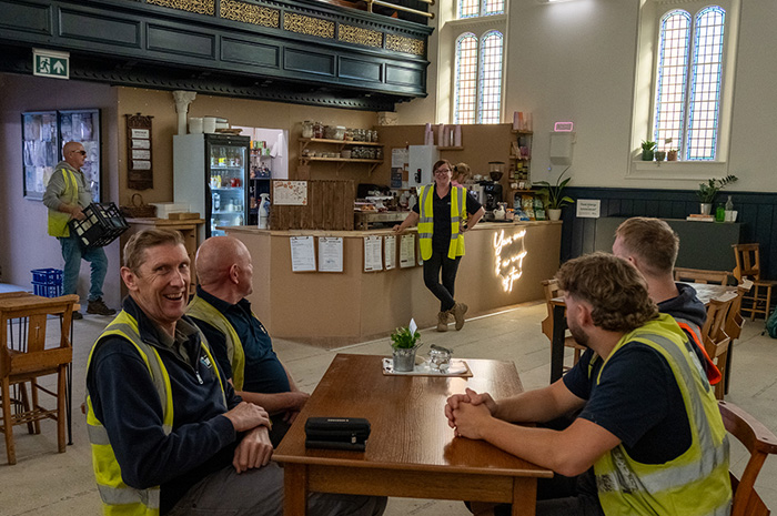 Workmen in hi viz vests drink coffee while Hayley stands behind them