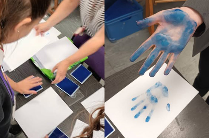 Looking over the shoulder of a pupil as she makes a blue handprint with inkpads and white paper